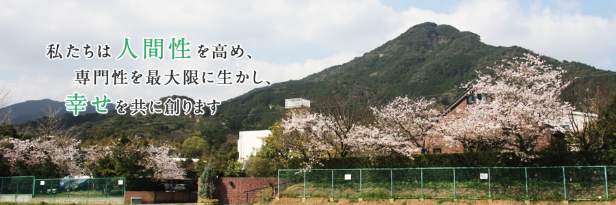 桜の木々と緑の山々の風景「私たちは人間性を高め、専門性を最大限に生かし、幸せを共に創ります」